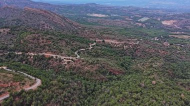 Yeşil tepeler, ağaçların arasından geçen virajlı yollarla araziye yayılıyor. Gökyüzü açık, gün boyunca İspanya kırsalında parlak bir manzara yaratıyor..