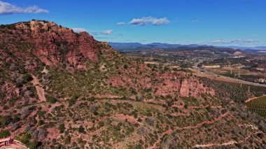 İspanya 'daki Sierra de Collserola dağlarının geniş bir manzarası kayalık tepeleri ve yeşil alanları gösteriyor. Açık mavi gökyüzünün altındaki tarlalar ve tarlalarla birlikte bir yol görülebilir..
