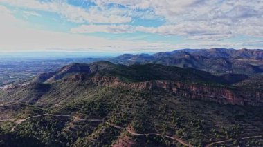 İspanya 'da mavi gökyüzünün altında yuvarlanan tepelerin ve vadilerin manzarası. Yeşil ağaçlar uzak mesafeden görülebilen kayalık oluşumlarla karayı kaplıyor. Öğleden sonra güneş parlıyor..