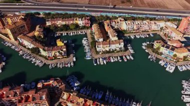 Tekneler marinada dizilmiş, deniz kenarında binalar var. Limana paralel giden bir otoyol var. Bu sahne İspanya 'da gün ışığında geçiyor..