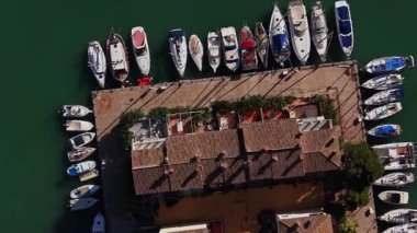 İspanya 'da ahşap bir rıhtım boyunca birkaç tekne park halindedir. Su berrak ve yeşil. Yakınlardaki binalar açık hava oturma yerlerini gösteriyor. Güneş sahne üzerinde ışıldıyor..