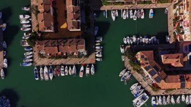 İspanya 'da bir marina manzarası rıhtım boyunca park etmiş birçok tekneyi gösteriyor. İnsanlar patika boyunca yürür. Binalar sahile yakın. Güneş sahnede parlıyor..