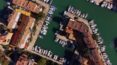 Gemiler marinadaki kanal boyunca dizildi. Suyun etrafındaki binalar ve güneş parlıyor. Bölgede insanlar yürüyor ya da dinleniyor olabilir..