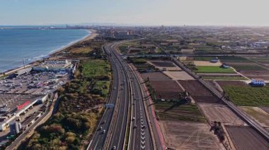 İspanya 'da kıyı boyunca uzanan bir otoyol var. Sahne iki taraftaki tarlaları ve uzaktaki okyanusu gösteriyor. Arabalar yol boyunca yol alırken güneş tepemizde parlıyor..