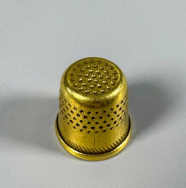 Altın bir yüksük hafif bir yüzeyde oturur. Yüksüğün dikiş için delikleri ve desenli bir tasarımı var. Durgun ve odadaki ışığı yansıtıyor..