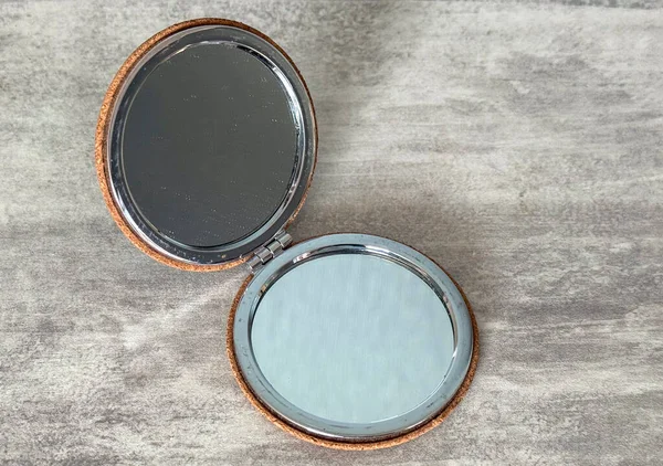 Deri kaplı yuvarlak bir ayna gri bir yüzeyde açık duruyor. Cam ışığı yansıtır ve net görüntüler ortaya çıkarır. Kişisel kullanım için basit bir aksesuar..