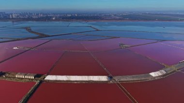 İşçiler su dolu bölümlere ayrılan tuz havuzlarını yönetirler. Manzara, yakınında tuz tepecikleri olan pembe ve mavi suları gösteriyor. Şehrin ufuk çizgisi görünür..