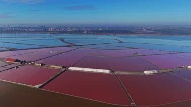 Tuz, kare şeklinde sıralanmış büyük göletlerde üretiliyor. Açık mavi gökyüzünün altında bir şehir silueti görülebilir..