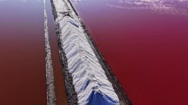 İşçiler renkli suların yakınındaki endüstriyel bir yerdeki büyük yığınlardan tuz topluyor. Bu sahne tuz üretiminin parlak güneş ışığı altında gerçekleştiğini gösteriyor..