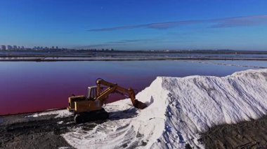 Bir makine suyun yakınındaki büyük bir kazıktan tuz taşır. Sahne pembe bir göl ve arka planda açık bir gökyüzünün altındaki binaları gösteriyor..
