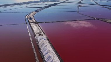 Parlak renklerde büyük tuz göletleri. İşçiler ve makineler sahnede görülebilir. Gökyüzü suyu yansıtıyor, eşsiz bir görünüm yaratıyor..