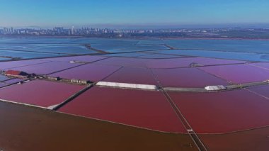 İşçiler gün içinde geniş tuz tavalarında tuz üretimini yönetirler. Şehir ufku renkli su birikintilerinin ötesinde görülebilir..