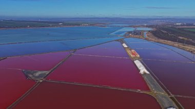 İşçiler, suyun dikdörtgen havuzlarda toplandığı tuz tarlalarını yönetirler. Suyun parlak renkleri, güneşin altında geniş bir arazide kontrasttır..