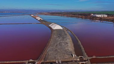 Tuz düzlükleri sığ havuzlarda canlı kırmızı su içeren büyük tuz yığınları sergiler. Sahne, kuru bir alanda tuz çıkarma sürecini yakalıyor..