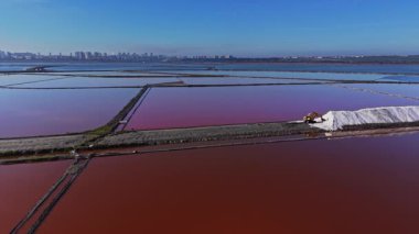İşçiler ve makineler bir tuz üretim alanında meşguller. Geniş tuz düzlükleri açık gökyüzünün altında çeşitli renklerde görünür. Yakın şehir silueti uzaklıktan görülebilir..