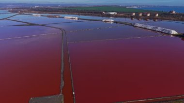 Tuz göletleri, güneş ışığını yansıtan ve farklı su renklerini gösteren bölümler halinde dizilmiştir. Bölge, arka planda binalar bulunan sahile yakın..