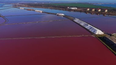 Tuz düzlükleri güneş altında parlak renklerle araziye yayılır. Tuz yığınları suyun yanında oturur ve tarlalarda desenler oluşturur. Binalar arka plan çizgisi.