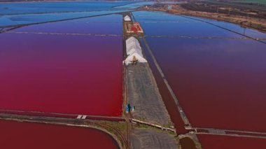 İşçiler parlak bir günde bir tuz çiftliğindeki renkli su havuzlarından tuz toplarlar. Manzara, güneşin tuzu kurutmak için kullandığı geniş düz alanlardan oluşur..
