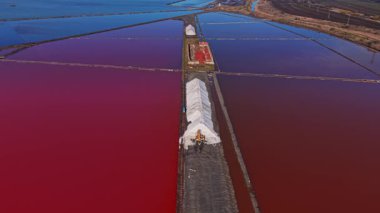 İşçiler, canlı pembe ve maviyi gösteren geniş tuz havuzlarından tuz toplarlar. Güneş suyun üzerinde parlarken insanlar tuzu toplamak için etrafta dolanıyor..