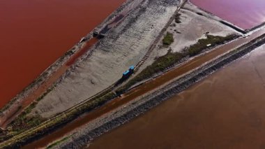 İşçiler kıyı bölgesindeki sığ göletlerden tuz topluyor. Kamyonların yol boyunca tuz madde taşıdığı görülüyor. Güneş sudan yansıyor..
