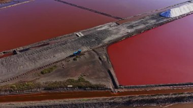 Tuz, suyun kırmızı bir renk yarattığı büyük tarlalarda toplanır. İşçiler karada kamyonlarla malzeme taşıyorlar. Yakınlarda tuz yığınları görülebilir..