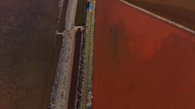 İşçiler kıyı boyunca bir tuz üretim alanında görülürler. Toprak, kırmızı ve kahverengi tonlarını gösteren renkli tuz göletlerine bölünmüştür..
