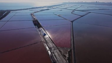 Tuz üretimi kıyıya yakın bir yerde gerçekleşir. Büyük dikdörtgen su birikintileri güneş ışığını yansıtır. Arka planda şehrin ufuk çizgisi görünür..