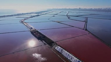 İşçiler güneş ışığı altında tuz tarlalarını yönetirler. Suyla dolu geniş düz alanlar renkleri yansıtır. Yakın şehrin ufuk çizgisi tuz endüstrisine bağlam katıyor.