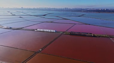 Tuz tarlaları değişik renklerde araziye yayılır. Açık mavi gökyüzünün altında bir şehrin ufuk çizgisi görünür..