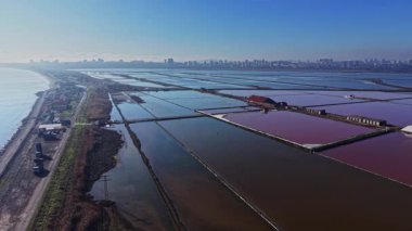 Tuz üretim alanları açık bir gökyüzünün altında arazi boyunca uzanıyor. Su, yapıların etrafında farklı tonlarda görülebilir. Şehir silüeti çok uzakta..