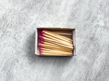 Küçük bir kutuda içinde pembe uçlu birkaç ahşap kibrit var. Kutu gri bir masanın üzerinde duruyor. Kibritler ateş yakmak için hazır..