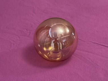 Yuvarlak cam bir nesne pembe bir yüzeye oturur. Cisim bakır rengine sahiptir ve ışık yansımalarını gösterir. Yumuşak ışıklandırma şeklini artırdığı için ayarlar basit..