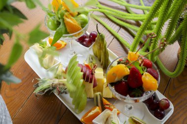 Meyve tabağında kaselerde çeşitli taze meyveler yer alır. Üzüm, portakal, çilek ve kivi zevk için ahşap bir masada sergilenir..
