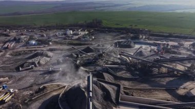 İşçiler taş ocağından malzeme alıyorlar. Ağır makineler kullanımda ve kamyonlar kaynakları taşıyor. Manzara açık araziyi ve çeşitli ekipmanları gösteriyor..