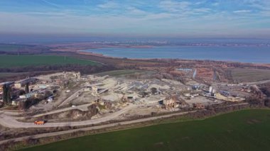 Görüntü aktif madencilik yapılan bir taş ocağı gösteriyor. Ağır ekipmanlar taşların ve toprağın arasında çalışıyor. Yakınlarda, arka planda bir su kütlesi ve yeşil alanlar var..
