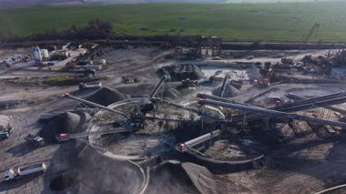 Ağır makineler bir taş ocağında çalışıyor. Taşıyıcı bantlar malzemeyi arazide hareket ettirir. Kamyonlar yoğun bir sanayi bölgesinde gün ışığında ağır yükleme yapmak için bekliyorlar..
