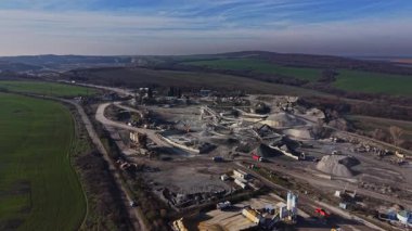 Kırsal alanda bir taş ocağı operasyonu başlatıldı. Kamyonlar park halindeyken ağır makineler malzemeleri taşır. Mavi gökyüzünün altında toprak ve taş yığınları görülebilir..