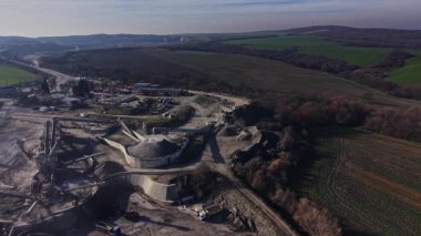 İşçiler taş ocağında makine kullanırlar. Ekipman, kamyonlar yakınlarda park halindeyken malzemeleri taşıyor. Manzara, berrak bir gökyüzünün altındaki tepeleri ve tarlaları gösteriyor..