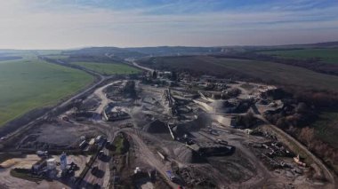 İşçiler ve makineler büyük bir taş ocağında taş ve mineralleri aktif olarak kaldırıyorlar. Çevrede yeşil tarlalar ve tepeler yer alırken, madencilik ekipmanları bölgede görülebiliyor..