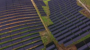 Sahne, açık gökyüzünün altındaki geniş yeşil arazi boyunca düz çizgiler halinde dizilmiş birçok güneş panelini gösteriyor. Bu sistem güneş enerjisi üretir ve yenilenebilir enerji sağlar..