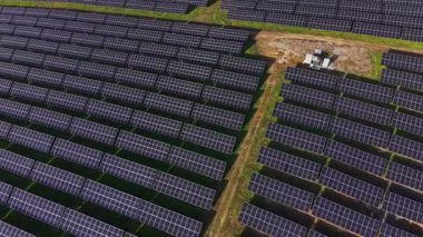 Güneş panelleri zemin boyunca sırayla dizilir. Güneş tepemizde parlıyor ve işçiler güneş çiftliği sahasının merkezinde küçük bir aracın yanında mevcutlar..