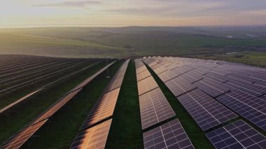 Güneş enerjisi panelleri yamaçta dizilmiş. Güneş batıyor, panellere yumuşak ışık saçıyor. Manzaranın arka planında yeşil tarlalar ve uzak tepeler vardır..