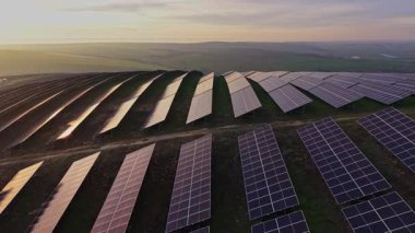 Güneş panelleri, güneş batarken tepedeki düzgün sıralara dizilir. Sahne uçsuz bucaksız yeşil bir alanı ve mavi gökyüzünü gösteriyor. Bu bölgede enerji üretimi yapılıyor.