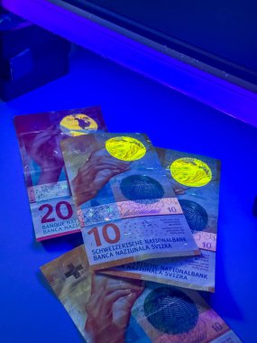 Mavi ışık altında parlayan bir yüzeye çok sayıda İsviçre frangı yerleştirilir. Banknotlar farklı değerleri gösteriyor ve eşsiz güvenlik özelliklerini açıkça vurguluyor..
