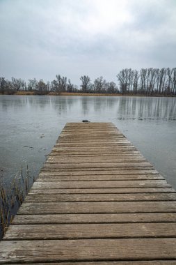 Yıpranmış ahşap bir iskele kısmen donmuş bir göle uzanır gri, bulutlu bir gökyüzünün altında. Çıplak ağaçlar uzak kıyı boyunca uzanır, buzlu suya hafifçe yansıyarak..