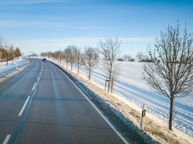 Dolambaçlı bir yol, berrak mavi bir gökyüzünün altında, karla kaplı bir kış manzarasını kesiyor. Çırılçıplak ağaçlar yol kenarında, uzak bir arabaya ve trafik ışıklarına doğru gidiyor..