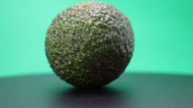 Bu büyüleyici görüntüde, bir avokado parlak siyah bir yüzey üzerinde zarifçe döner, canlı yeşil arkaplan ile çarpıcı bir kontrast yaratır.