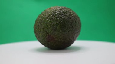 Tek bir olgun avokado yumuşak beyaz bir yüzey üzerinde nazikçe yuvarlanarak zengin dokusunu ve koyu yeşil rengini vurgular..