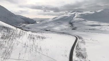 İnsansız hava aracı, öğleden sonra güneş ışığı altında dağların yamaçlarında karlar ve çıplak ağaçlarla çevrili kırsal bir yolda yavaşça ileri doğru uçuyor. Sakin ve sinematik İskandinav atmosferi. 