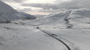 İnsansız hava aracı kar ve dağlarla çevrili kırsal bir yoldan yavaşça sola doğru uçuyor. Yumuşak akşam ışığı ve arka planda bulutlar barışçıl bir İskandinav bahar manzarası yaratır..